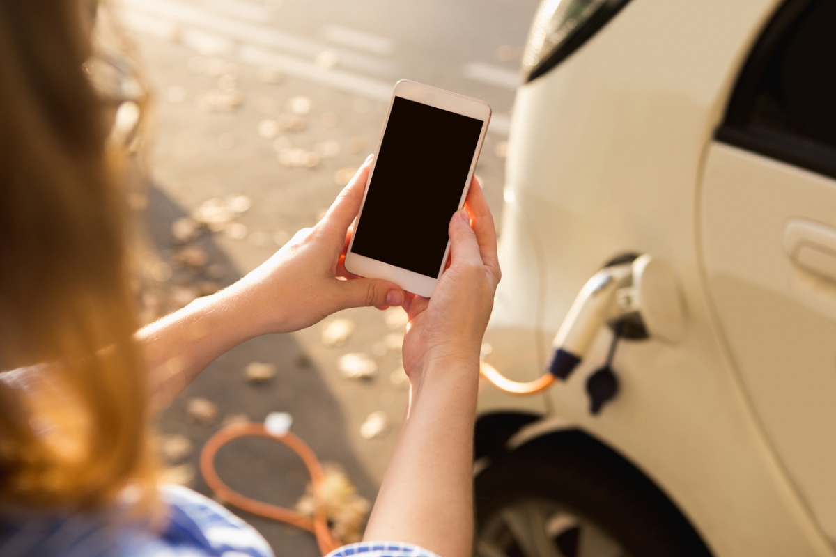 Junge Frau steht neben Mietauto, dass an Ladestation lädt und schaut auf Smartphone.