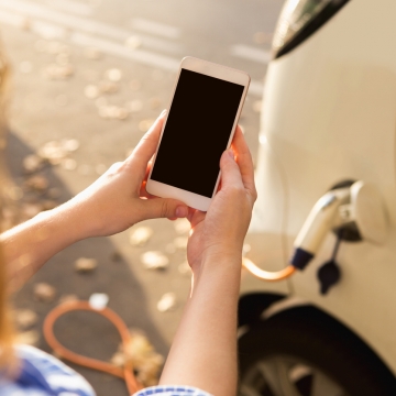 Junge Frau steht neben Mietauto, dass an Ladestation lädt und schaut auf Smartphone.